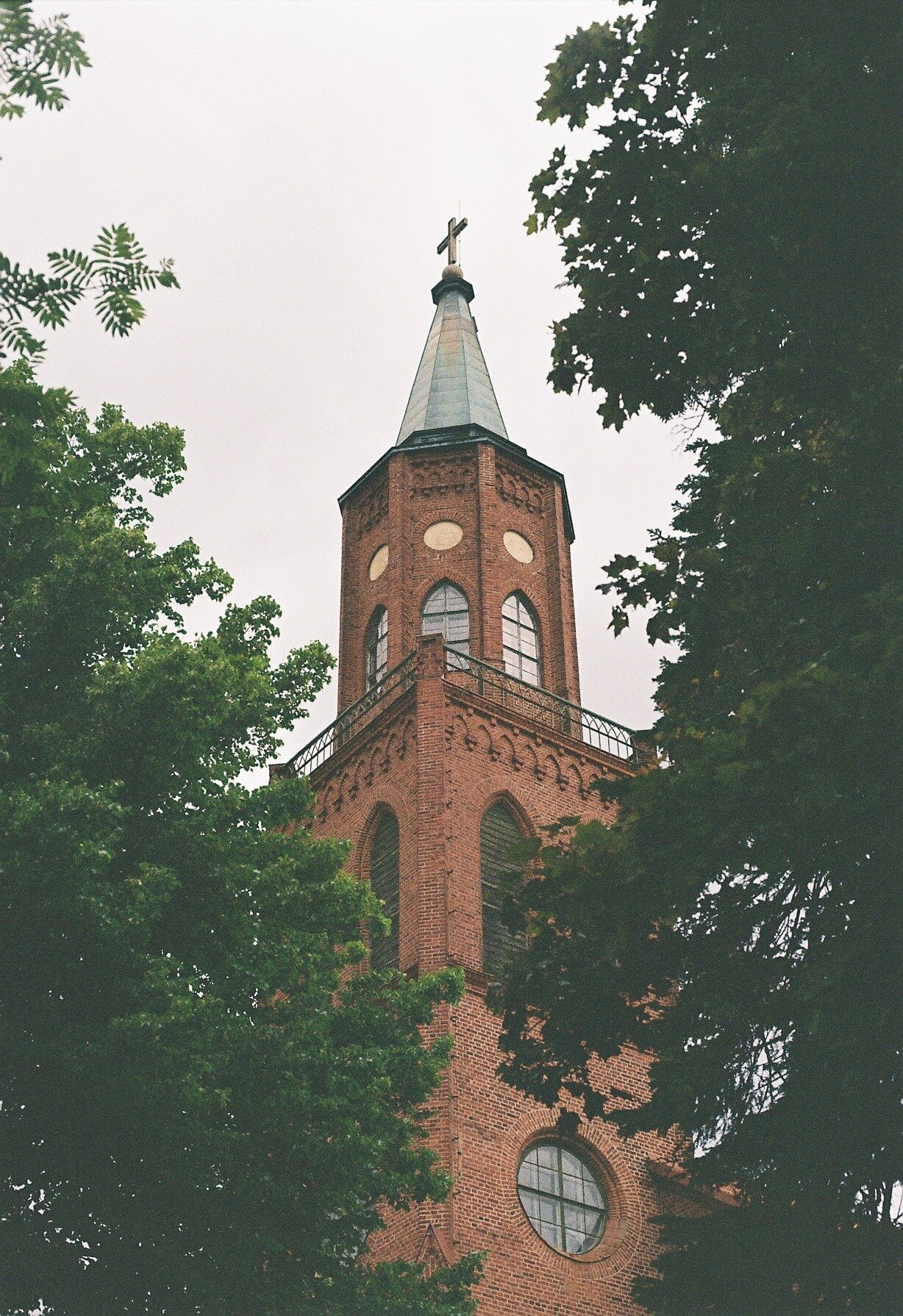 A red-brick church tower with a cross-topped spire, framed by leafy tree branches.