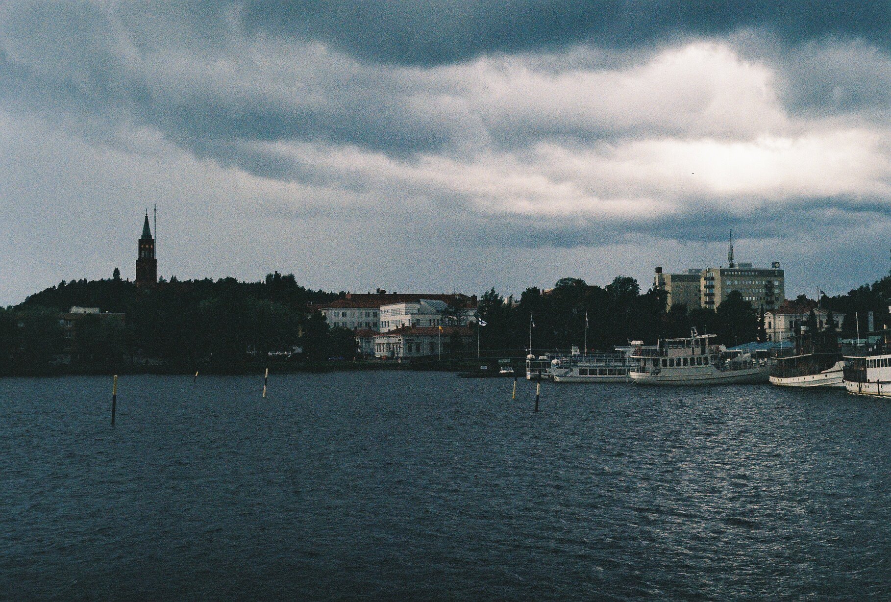 Old passenger boats moored at a harbor at dusk, a church spire silhouetted against dramatic storm clouds.