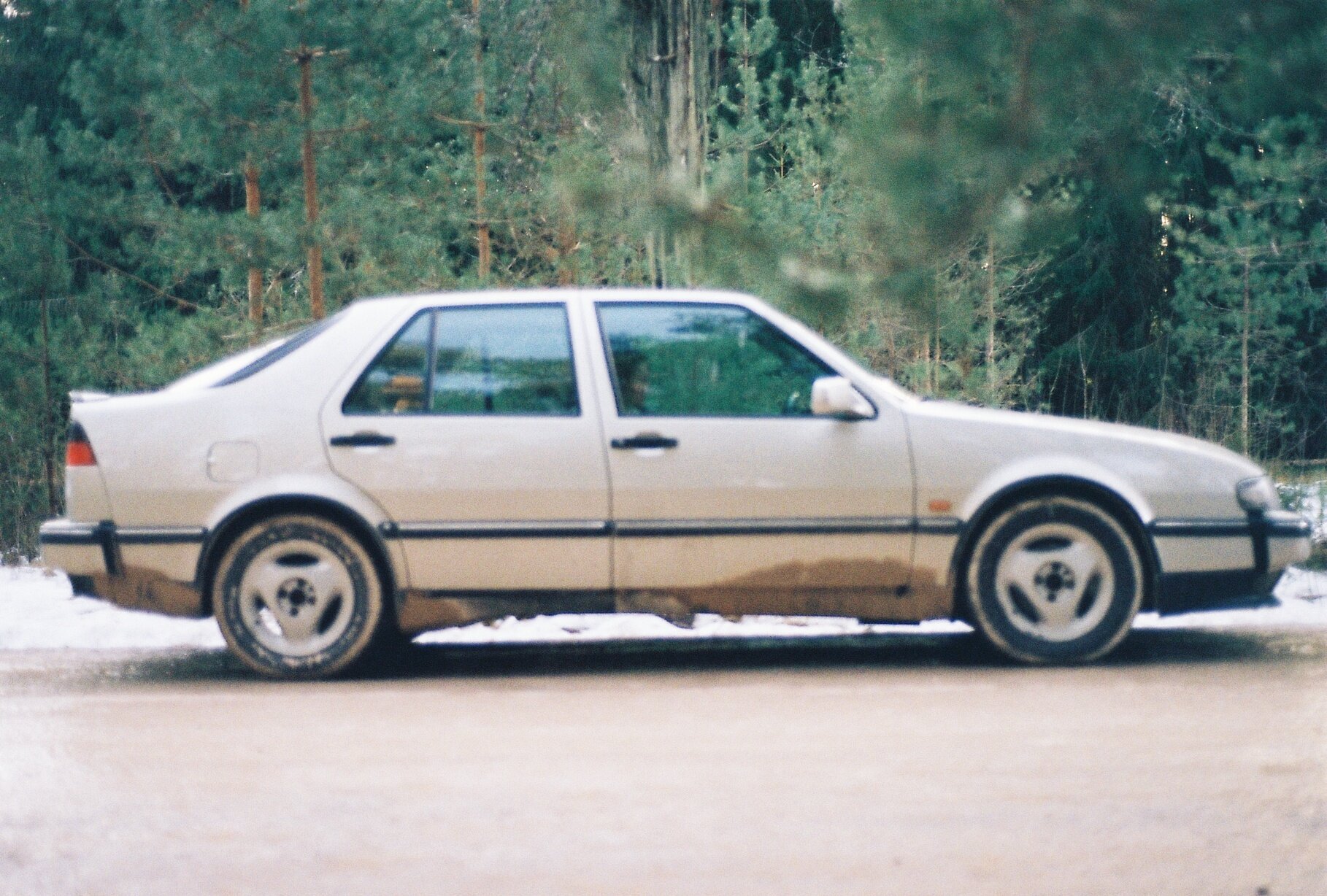 A silver Saab sedan parked on a forest road with patches of snow on the ground.