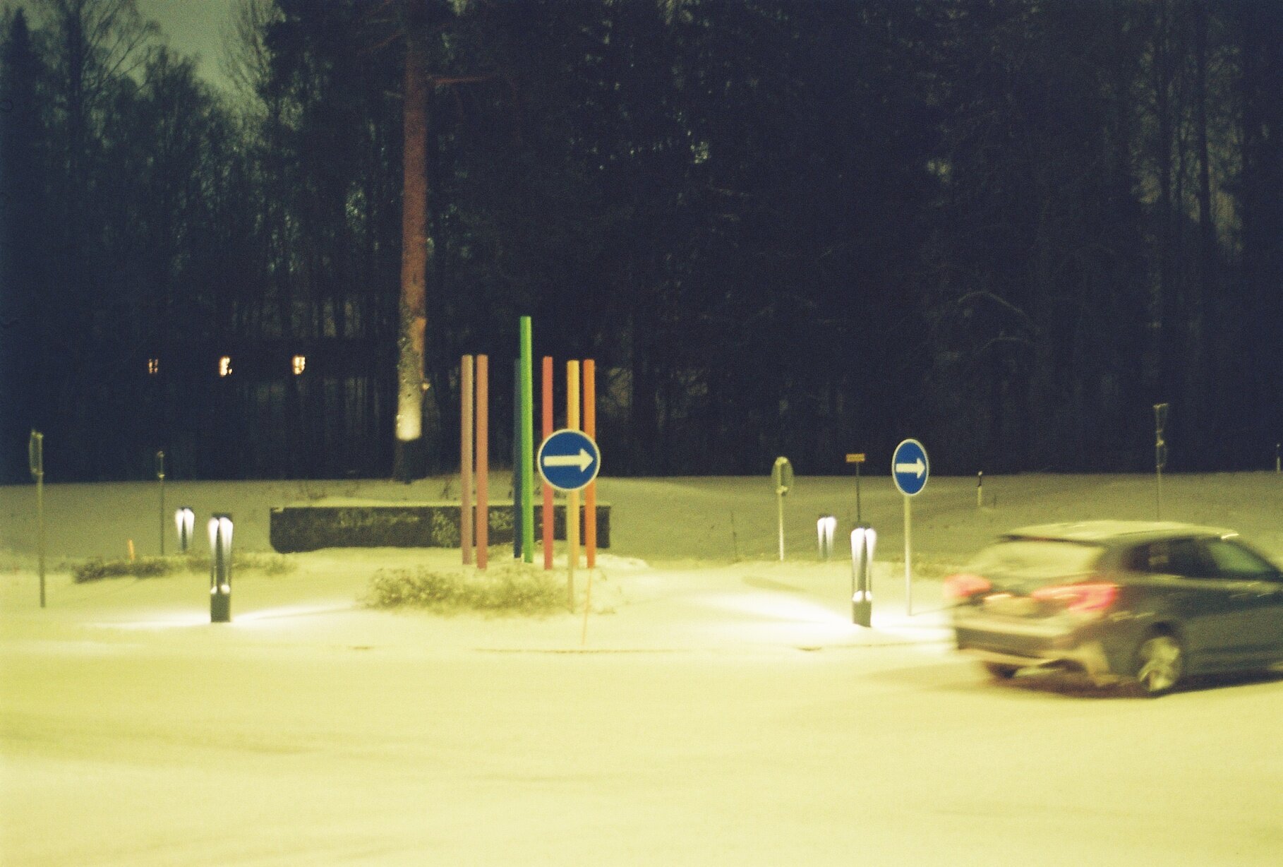 A snowy crossroads at night, colorful light poles and a one-way sign beside a passing car, forest in the background.